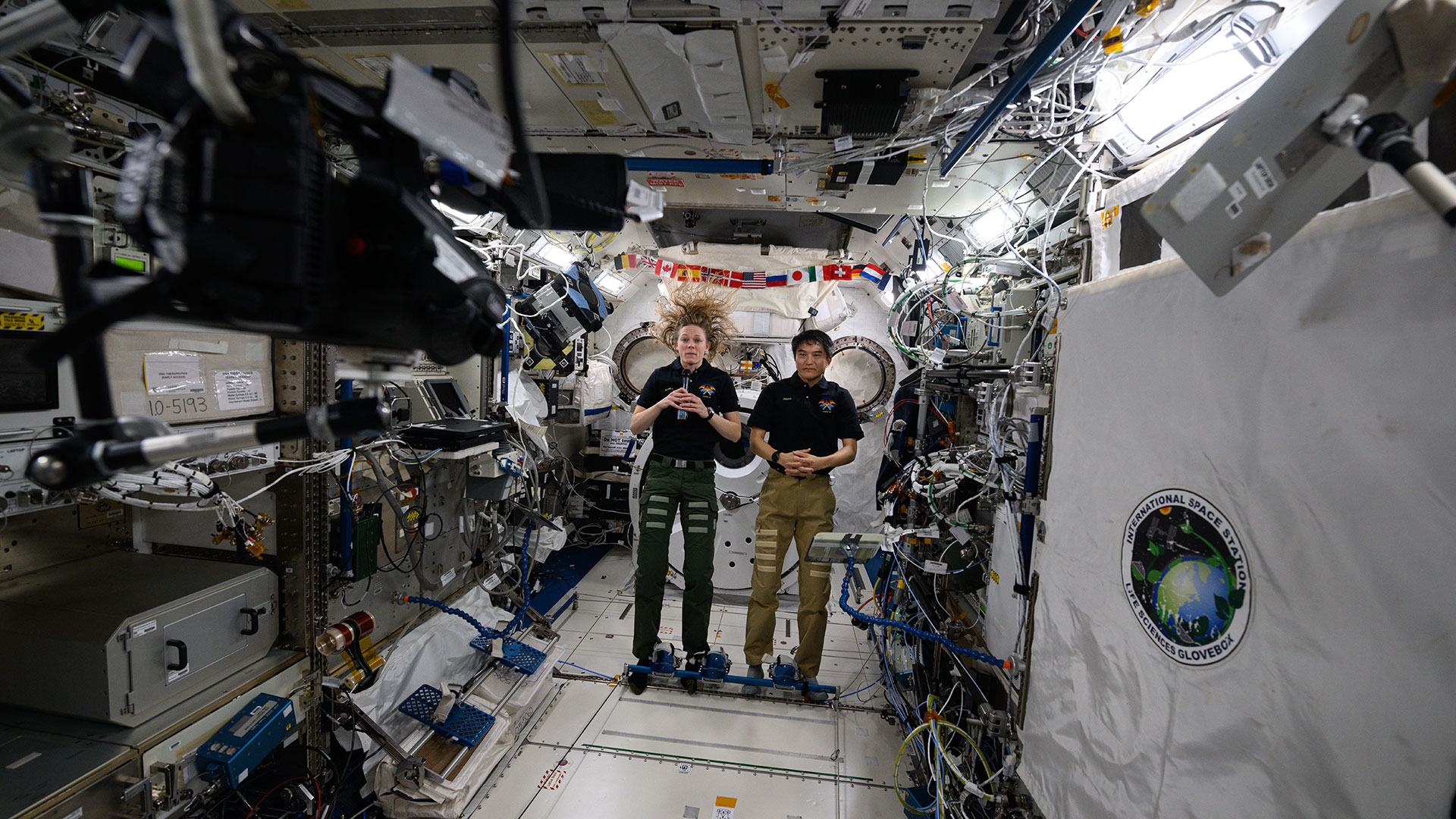 a woman and man, both wearing a polo shirt and slacks, speak to a camera aboard a space station